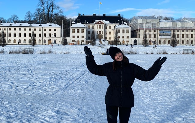 En kvinna i svart kappa poserar för foto i ett snöigt landskap med Karlbergs slott i bakgrunden.