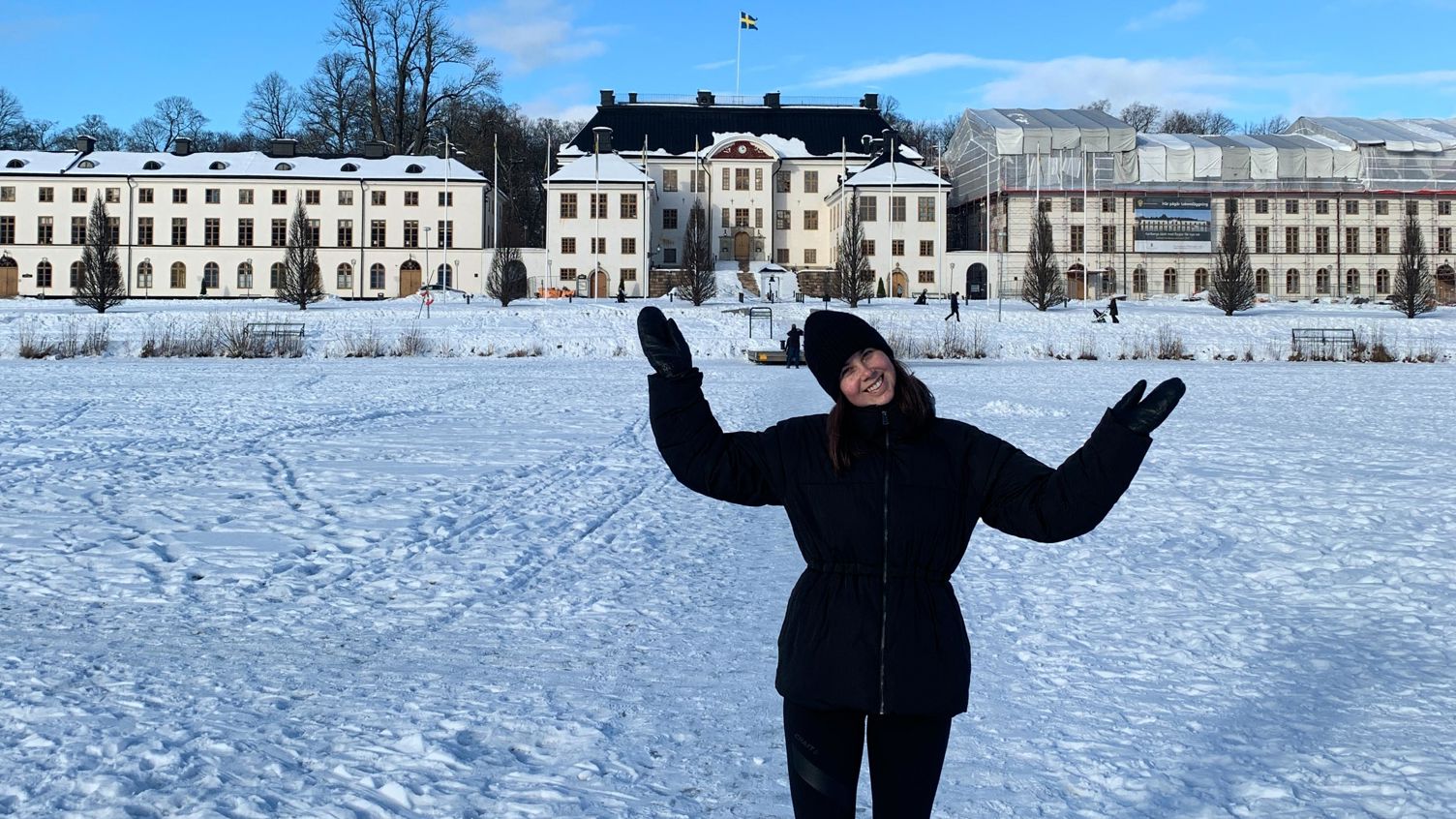 En kvinna i svart kappa poserar för foto i ett snöigt landskap med Karlbergs slott i bakgrunden.