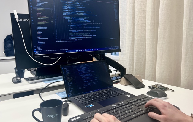 Närbild på en laptop och datorskärm som visar kod. En kopp med texten Zington på står bredvid.