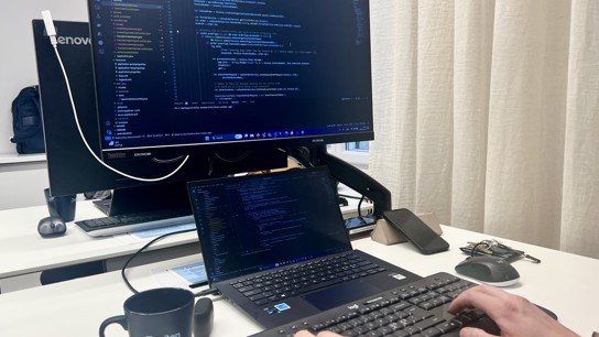 Närbild på en laptop och datorskärm som visar kod. En kopp med texten Zington på står bredvid.