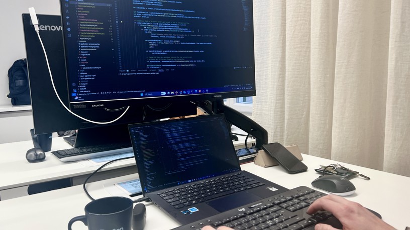 Närbild på en laptop och datorskärm som visar kod. En kopp med texten Zington på står bredvid.