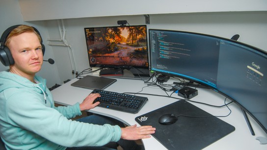 En man sitter med hörlurar vid ett skrivbord med två bildskärmar varav en är en panoramaskärm.