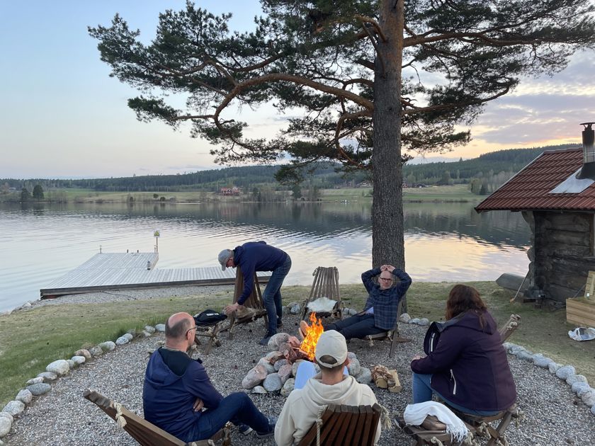 En grupp människor som sitter runt en eld med en sjö i bakgrunden.