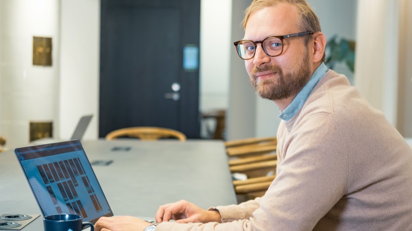 En man som sitter vid ett bord med en bärbar dator och kollar åt sidan mot kameran.