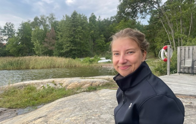Emelie Djenfer sitter på en klippa och ler, vatten och vass syns i bakgrunden.