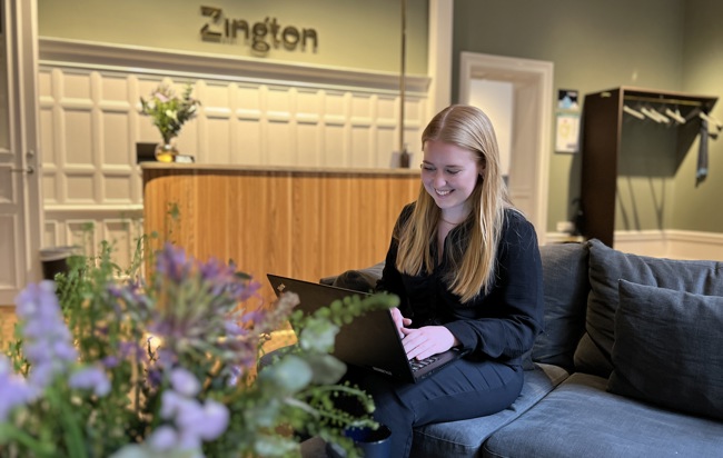 Zingtonmedarbetaren Melanie sitter i en soffa i Zingtons gröna lobby med en dator i famnen, hon ler.