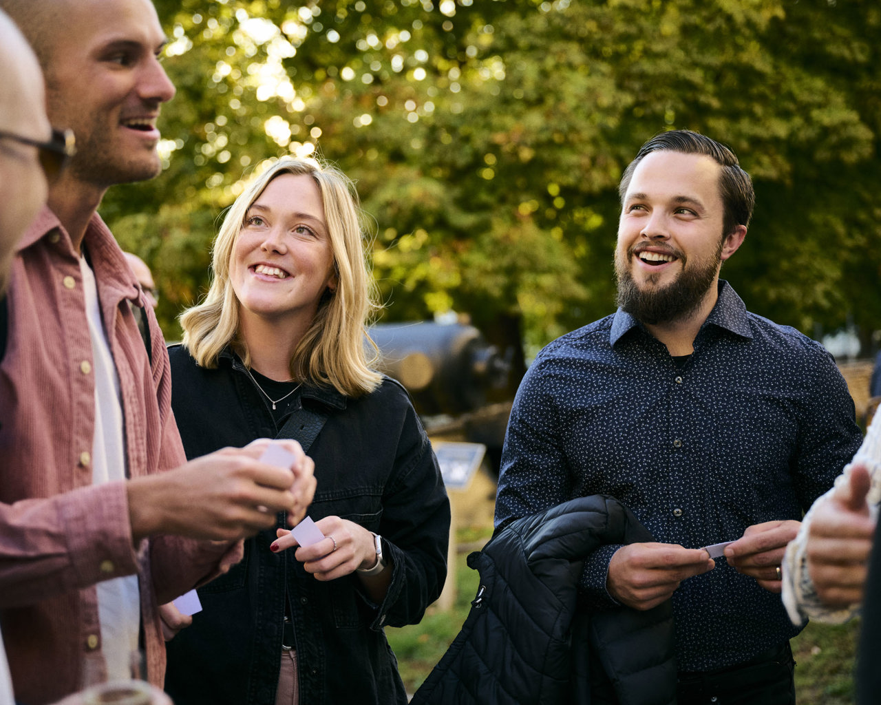 Vendela ler mot sina kollegor tillsammans med Jesper under en solig sensommarkväll, omgiven av en lummig utomhusmiljö.