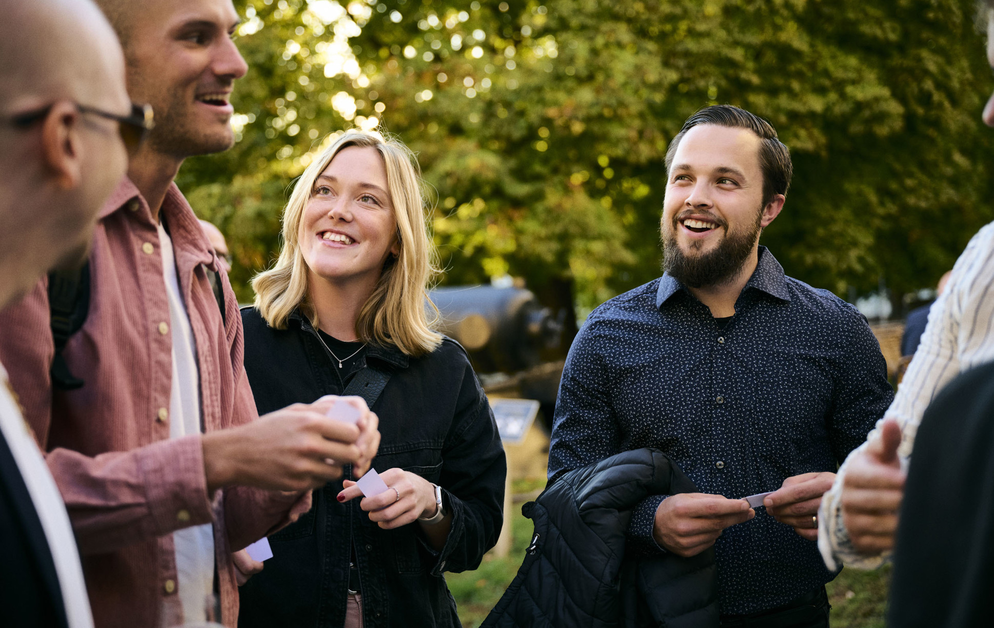 Vendela ler mot sina kollegor tillsammans med Jesper under en solig sensommarkväll, omgiven av en lummig utomhusmiljö.