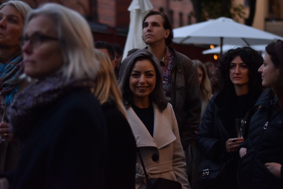 En grupp människor som står utomhus. Fokus på en kvinna i ljus kappa som tittar rakt fram.