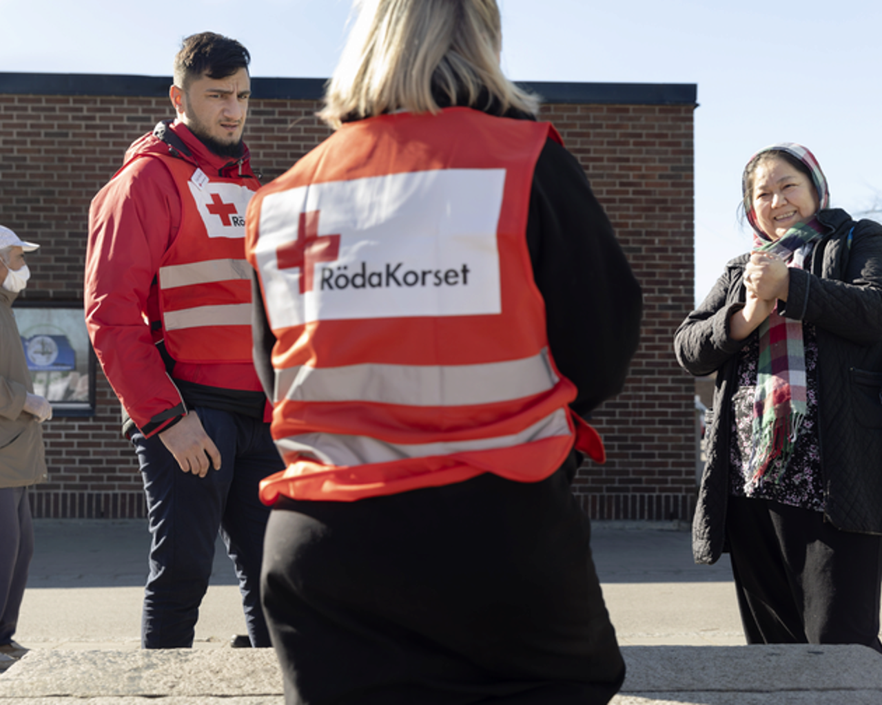 Två arbetare med röda korset västar pratar med en kvinna utomhus.