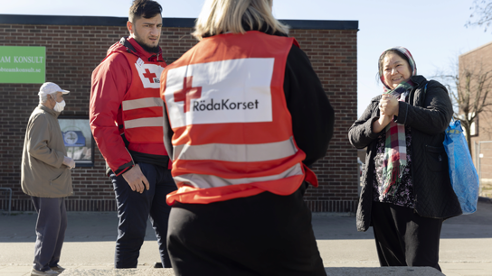 Två arbetare med röda korset västar pratar med en kvinna utomhus.