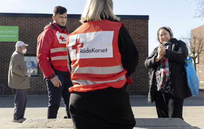 Två arbetare med röda korset västar pratar med en kvinna utomhus.