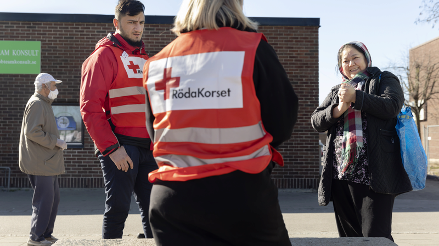 Två arbetare med röda korset västar pratar med en kvinna utomhus.