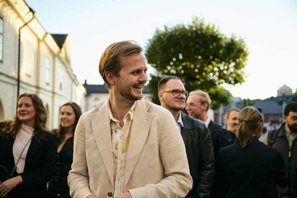 En man bland en grupp av människor i vit kavaj och kläder ler och kollar åt sidan utomhus.