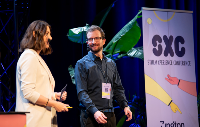 A woman and a man stand on a stage with a roll-up in the background with the text STHLM Xperience Conference.
