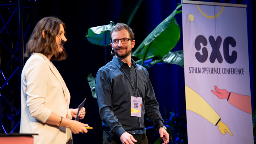 A woman and a man stand on a stage with a roll-up in the background with the text STHLM Xperience Conference.