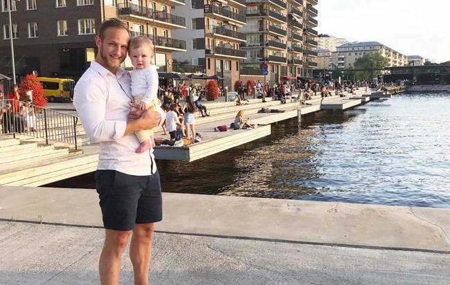 En man står med ett barn i famnen framför en kaj, omgiven av stadsmiljöns liv och rörelse.