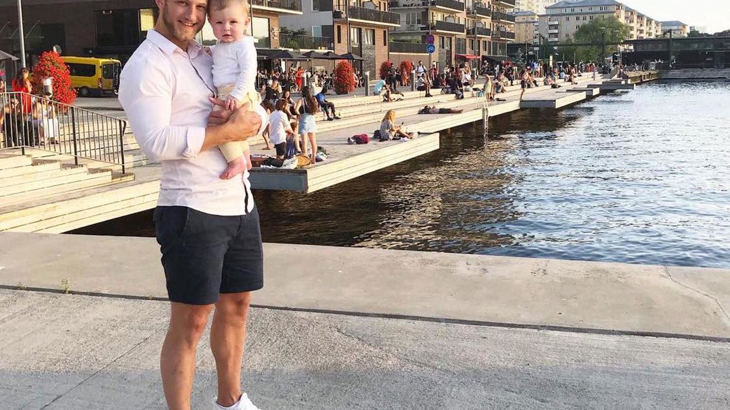 En man står med ett barn i famnen framför en kaj, omgiven av stadsmiljöns liv och rörelse.